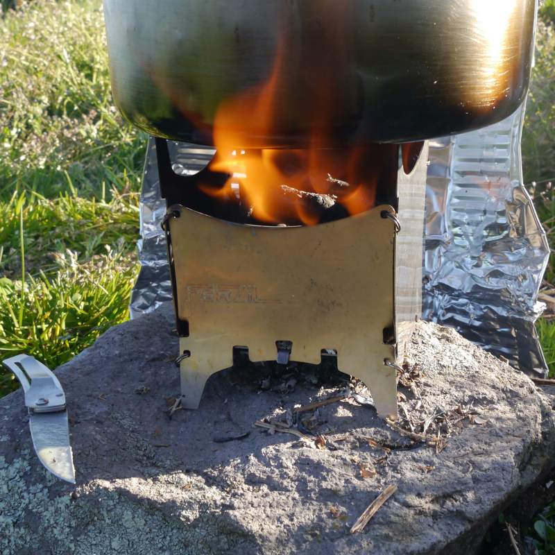 cuisine de bivouac, dehors au soleil, cuisine au feu de bois avec bushbox ferzil posé sur une pierre. Couteau. Paravent avec une barquette en aluminium